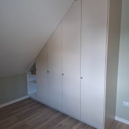 TOP PROJECT Piotr Dul - Jasnoszara szafa na wymiar z czterema drzwiami i otwartymi półkami, precyzyjnie wkomponowana w skos sufitu na poddaszu, z drewnianą podłogą i jasnymi ścianami.