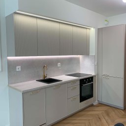 TOP PROJECT Piotr Dul - Nowoczesna kuchnia z ryflowanymi szafkami górnymi w odcieniu beżu, złotą armaturą, podświetlanym blatem i drewnianą podłogą w jodełkę.
