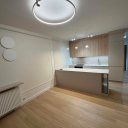 TOP PROJECT Piotr Dul - Nowoczesna otwarta przestrzeń z kuchnią w drewnie i beżu, wyspą z płytą grzewczą, oraz salonem z jasną podłogą, dekoracyjną listwą ścienną i okrągłą lampą sufitową.