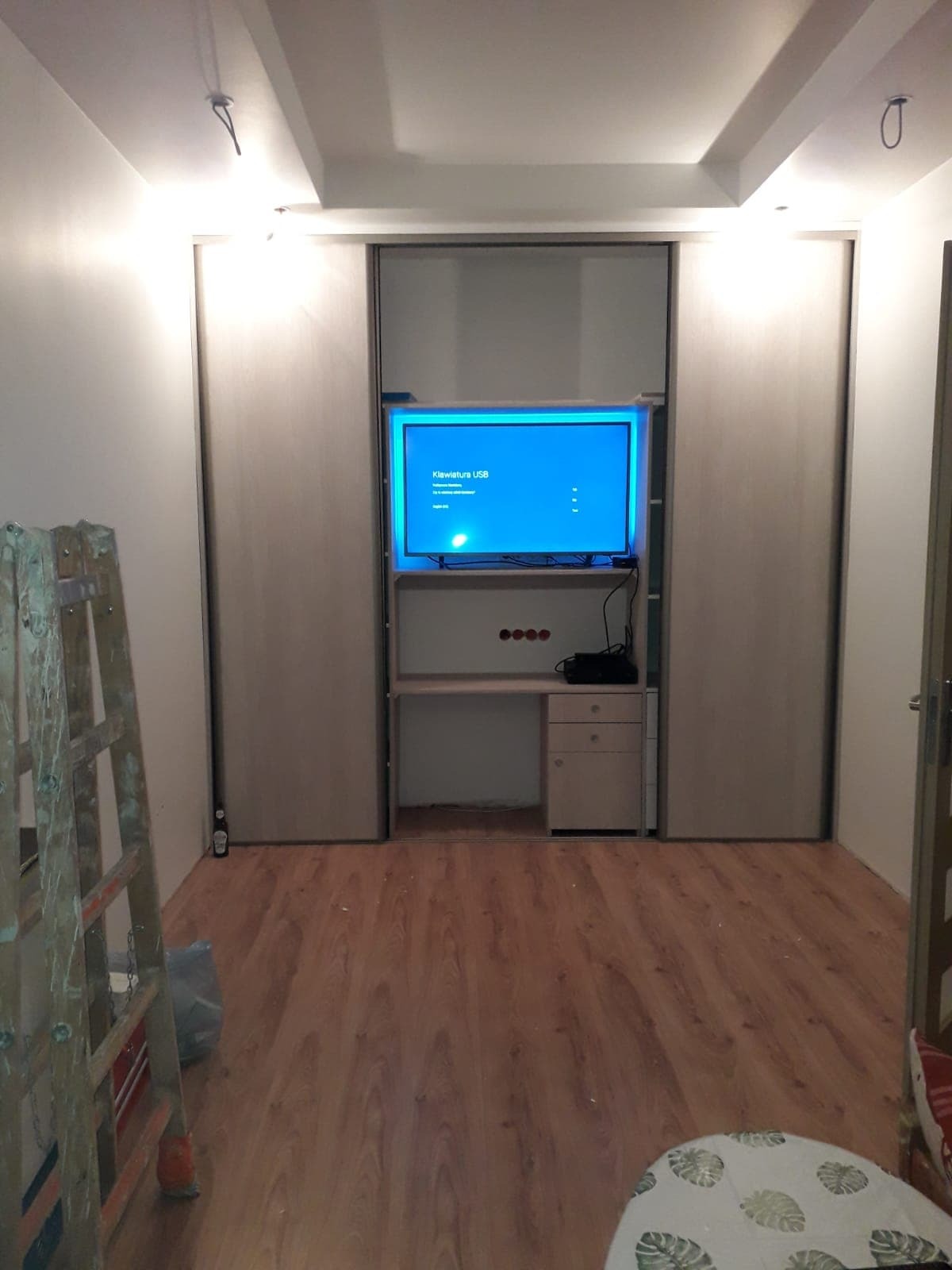 Wnęka z szafą przesuwną i biurkiem z telewizorem. Widoczna drabina malarska, sugerująca prace wykończeniowe. Podłoga z paneli. Oświetlenie sufitowe.