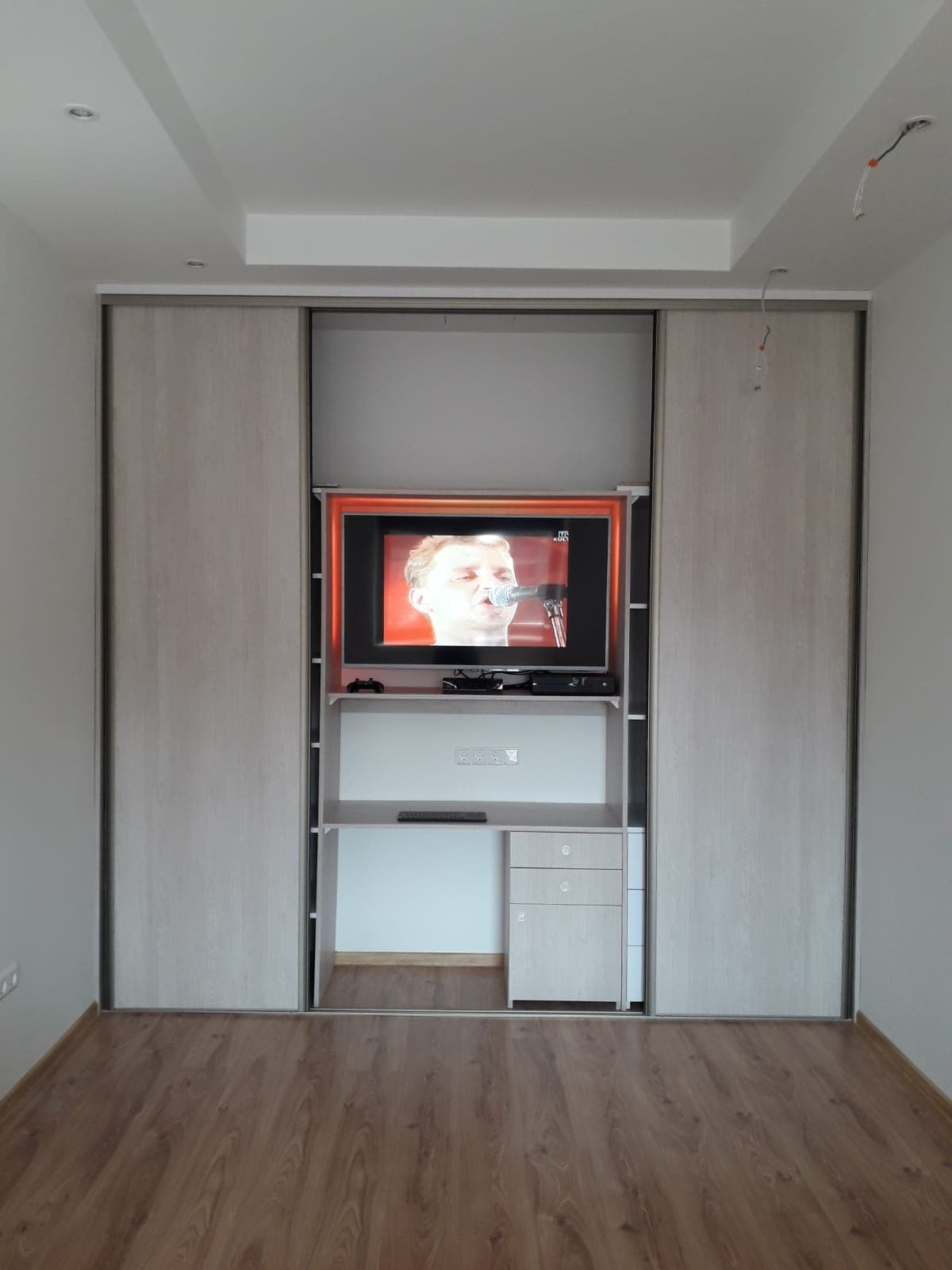 Szafa wnękowa z przesuwnymi drzwiami w odcieniach beżu, wkomponowana w ścianę, z wnęką na telewizor i biurko. Widoczna drewniana podłoga i fragment sufitu.