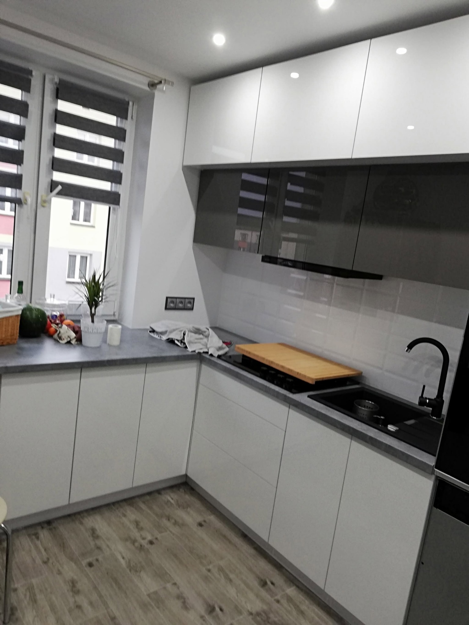 Nowoczesna kuchnia z białymi szafkami i szarym blatem, czarny zlew i bateria, drewniana deska na płycie. Widok na okno z roletami.