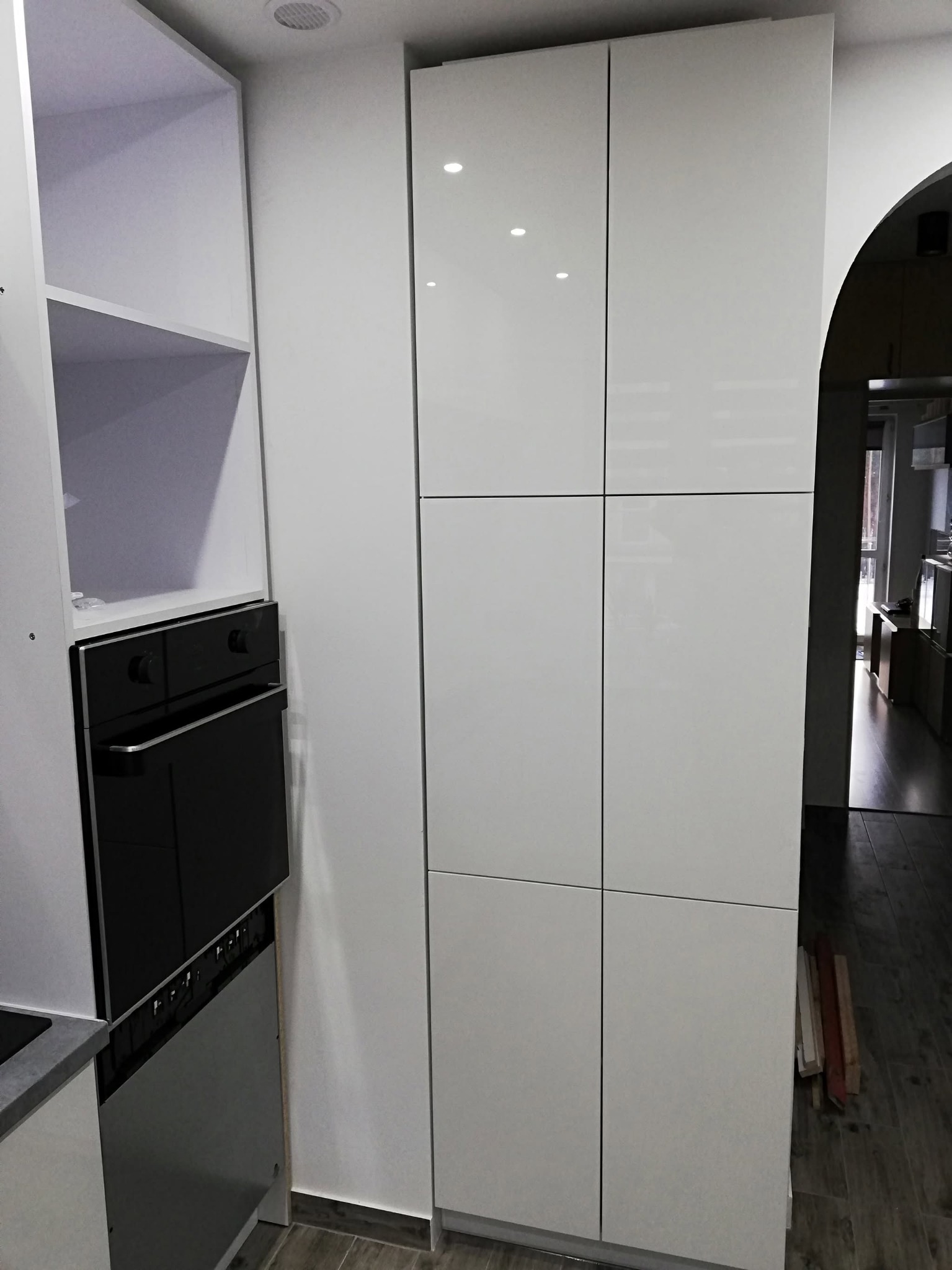 Biała, nowoczesna zabudowa kuchenna z piekarnikiem w trakcie montażu. Widoczne elementy szafek i frontów. Podłoga z ciemnego drewna. W tle widoczne inne pomieszczenie.