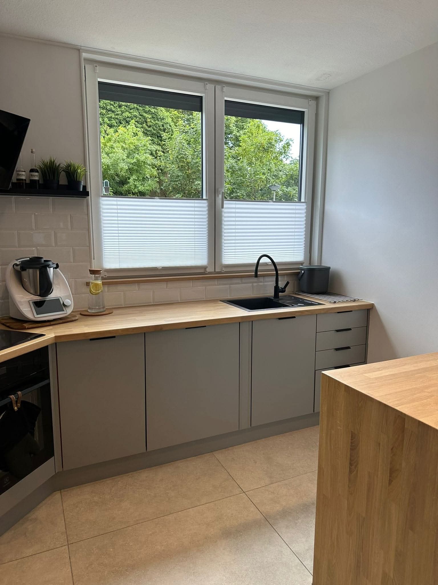 Szara zabudowa kuchenna z drewnianym blatem i czarną armaturą, widok na okno z żaluzjami plisowanymi. Na blacie robot kuchenny i akcesoria. Jasne płytki na podłodze.