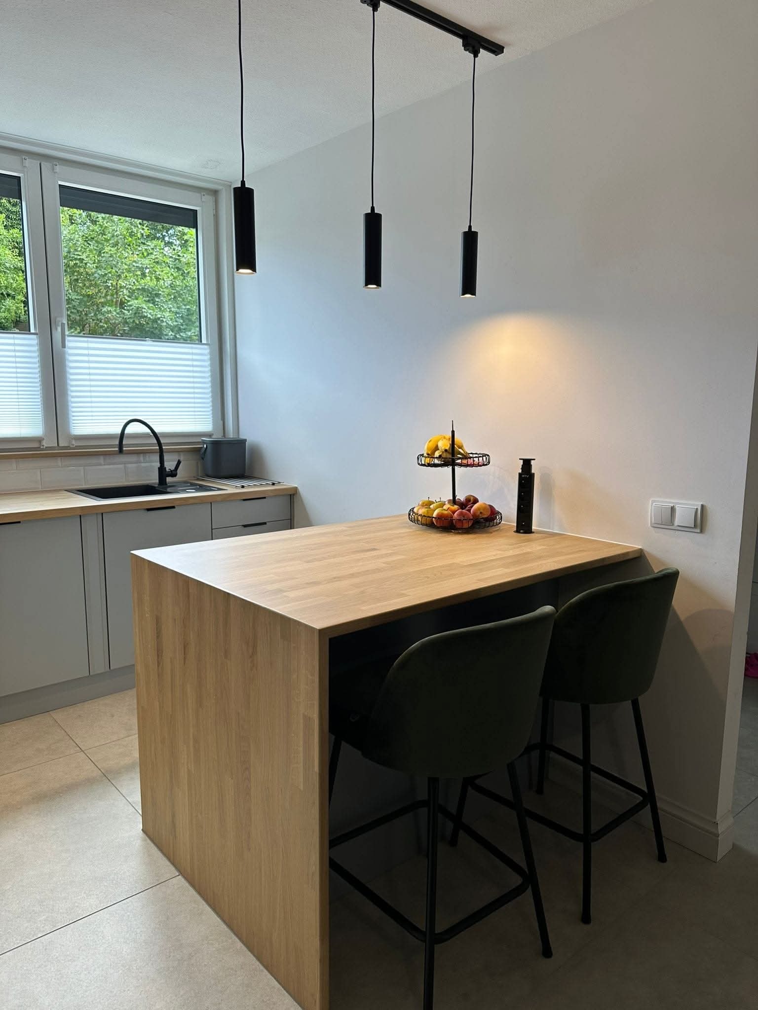 Nowoczesna kuchnia z wyspą o drewnianym blacie, oświetlona trzema cylindrycznymi lampami. Minimalistyczny design, szare szafki i krzesła barowe w kolorze butelkowej zieleni.