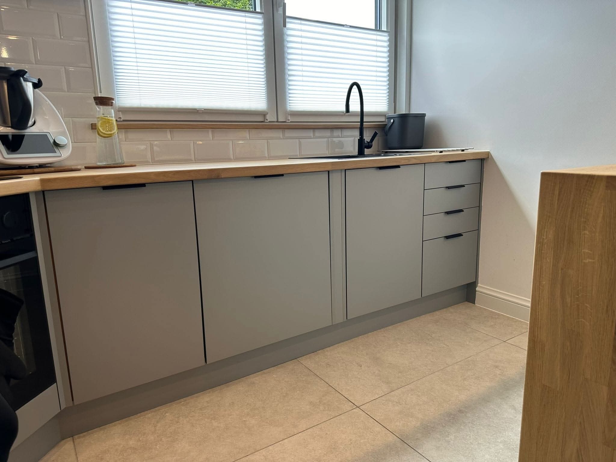 Szare szafki kuchenne z drewnianym blatem i czarną armaturą pod oknem z żaluzjami. Minimalistyczny design, jasne płytki podłogowe, nowoczesne wykończenie.