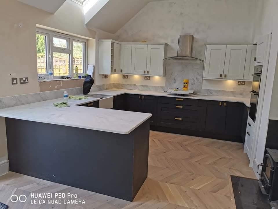 Nowoczesna kuchnia w odcieniach szarości i bieli z marmurowym blatem, parkietem w jodełkę i oknem dachowym. Widoczne detale wykończenia i oświetlenie podszafkowe.