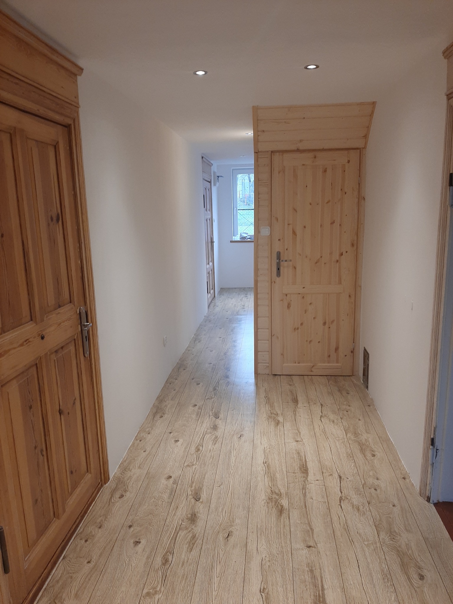 Wnętrze korytarza z nową podłogą imitującą drewno, widoczne dwie drewniane ościeżnice z drzwiami, białe ściany i sufit z oświetleniem punktowym.