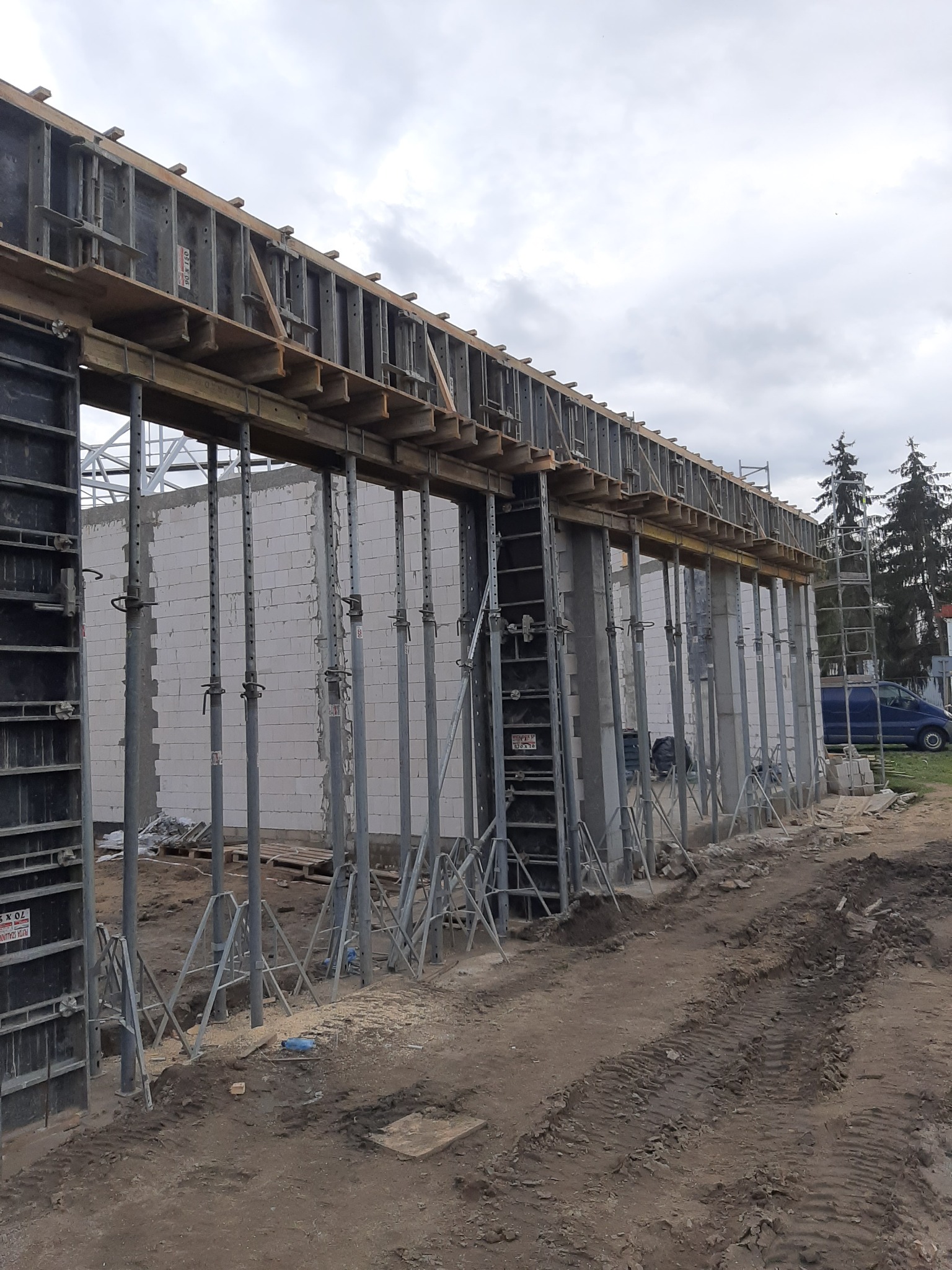 Szare, betonowe ściany budynku w trakcie budowy, wzmocnione metalowymi stemplami i drewnianymi szalunkami, na pierwszym planie ziemia z widocznymi śladami opon