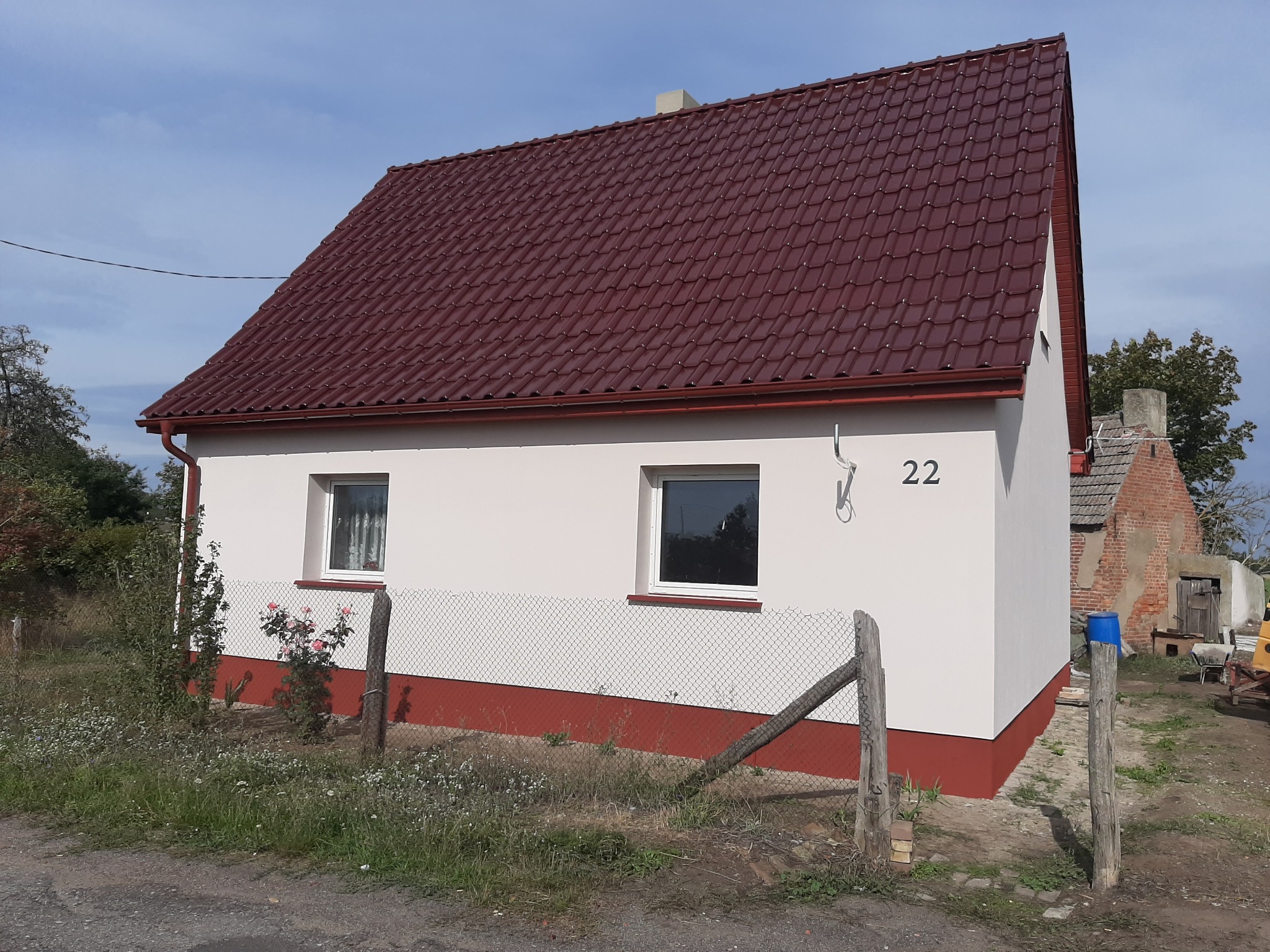 Odnowiony, parterowy dom z ciemnoczerwonym dachem, świeżo otynkowaną, jasną elewacją i numerem '22' na ścianie, widziany z perspektywy niskiego kąta.