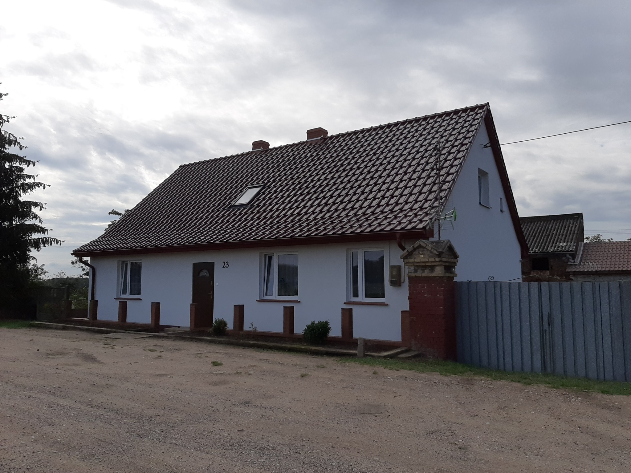 Biały dom jednorodzinny z brązową dachówką, widoczny numer domu 23, z odnowioną elewacją i częściowo odnowionym ogrodzeniem z ceglanymi elementami.
