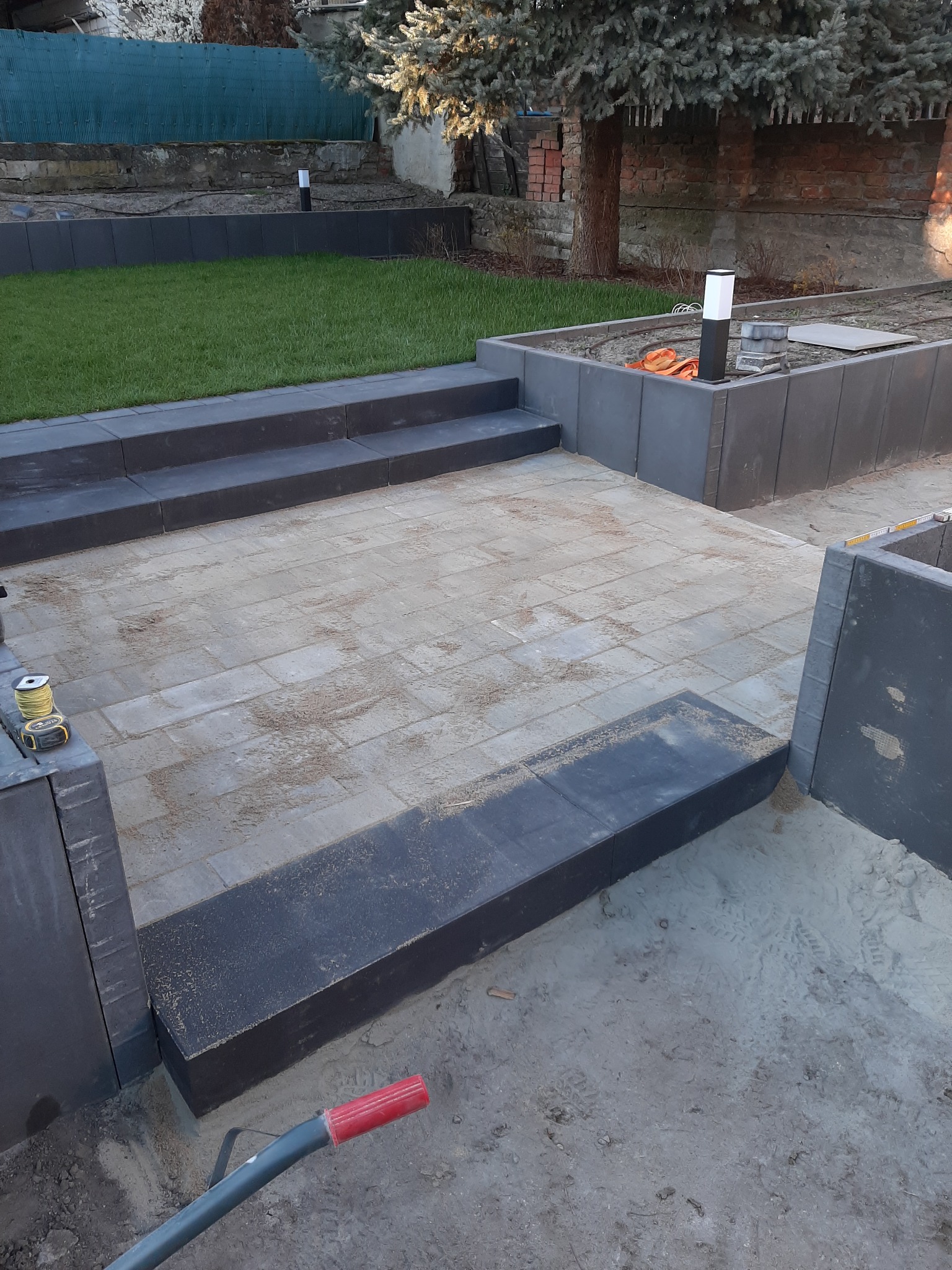 Układanie kostki brukowej na posesji z elementami dekoracyjnymi, widoczne schody i obrzeża z ciemnych płyt, trawnik w tle, częściowo ukończona praca.