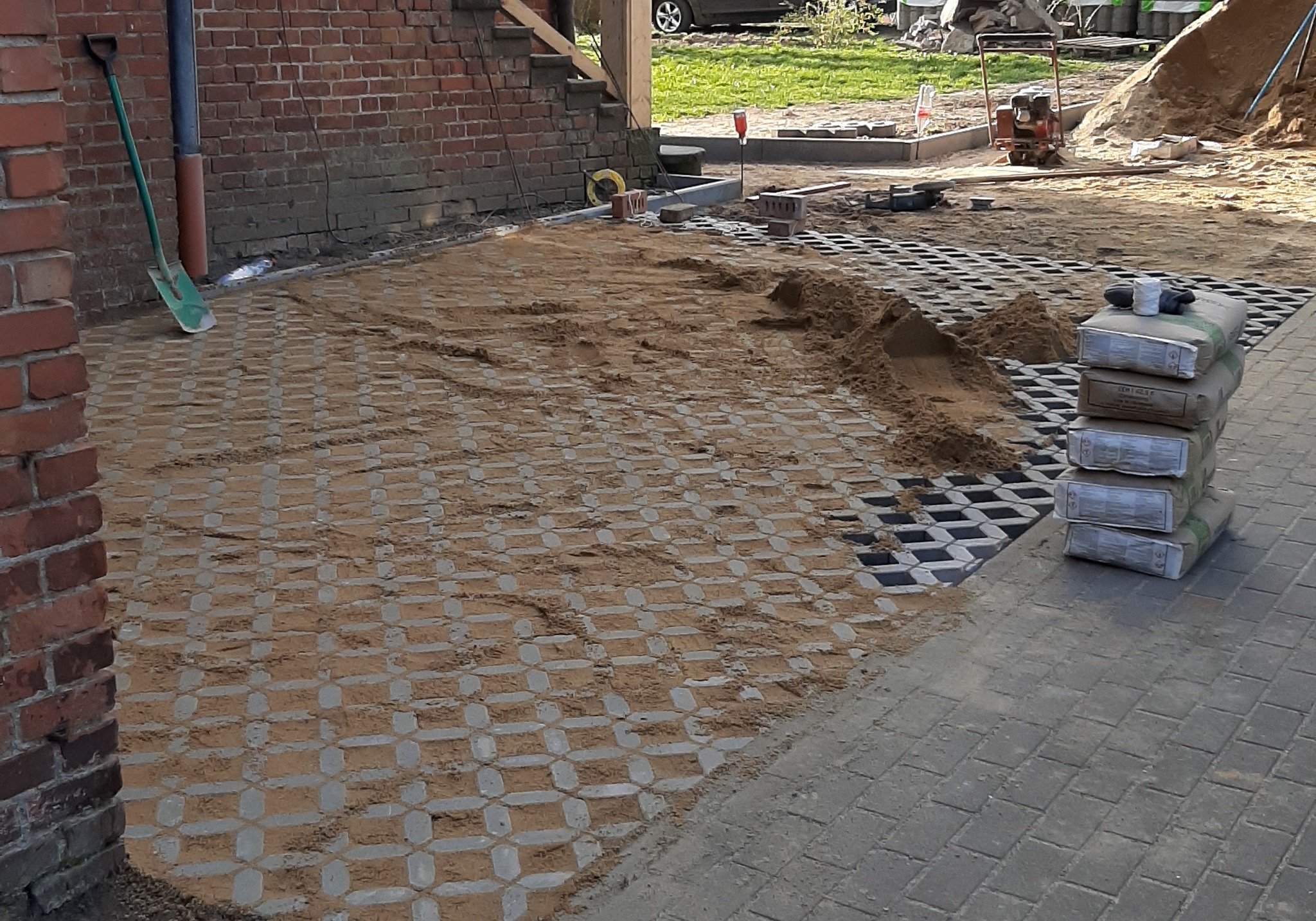 Przygotowanie podłoża pod nawierzchnię z ażurowych płyt betonowych; widoczne płyty wypełnione piaskiem, ubijarka w tle oraz paleta z workami materiału.