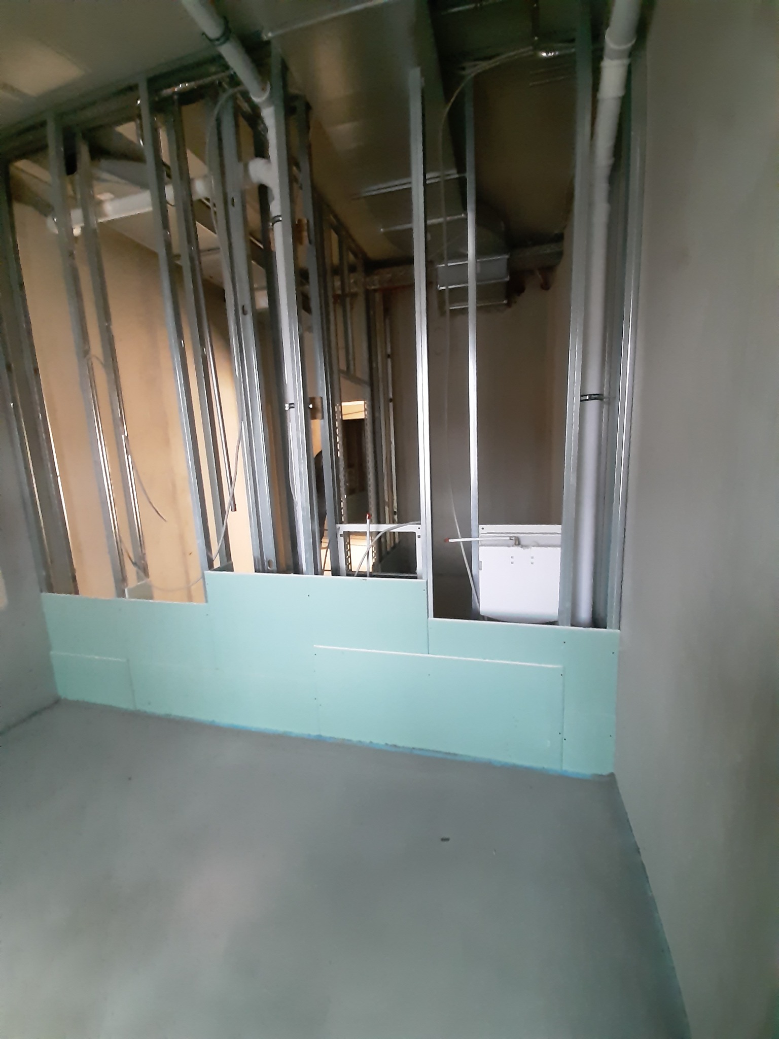 Wnętrze w trakcie montażu ścian działowych z metalowych profili i częściowo zamontowanych, zielonych płyt na dolnym poziomie. Widoczne instalacje w ścianach i surowy beton na podłodze.