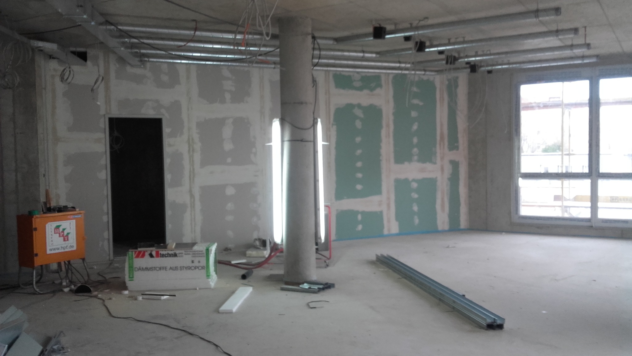 Wnętrze w trakcie prac wykończeniowych: ściany z płytami gipsowo-kartonowymi częściowo zaszpachlowane, widoczny słup konstrukcyjny z lampami, instalacje pod sufitem, materiały budowlane i narzędzia...