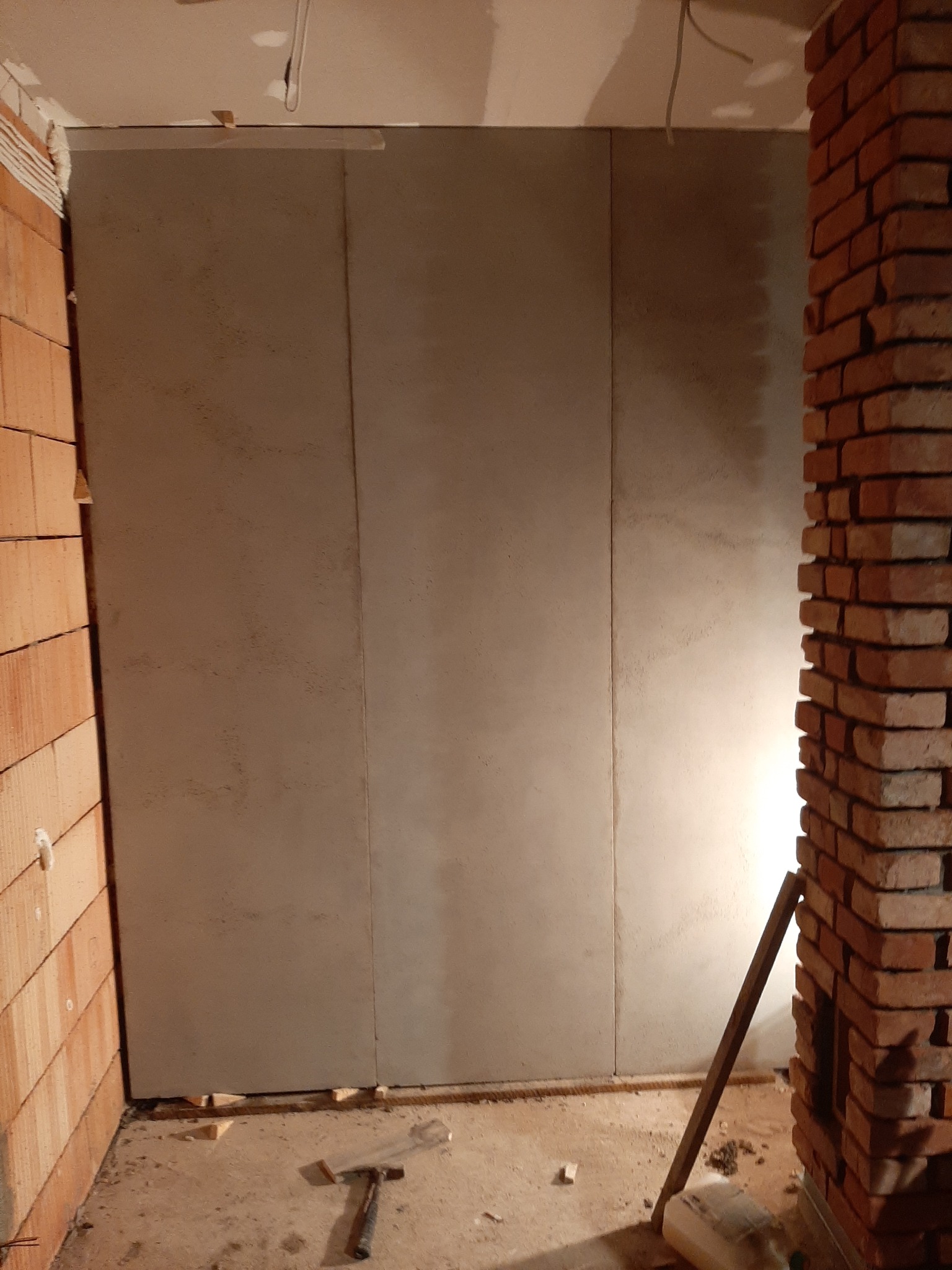 Wnętrze w trakcie prac wykończeniowych: ściana z płyt pokryta szpachlą, widoczne łączenia płyt, fragment ściany z cegły ceramicznej, młotek i inne narzędzia na podłodze.
