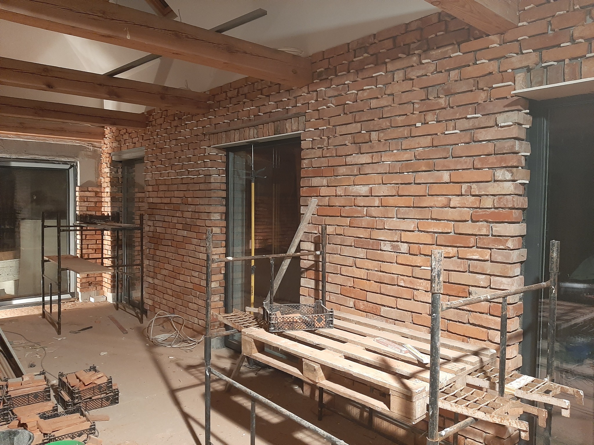 Wnętrze w trakcie wykańczania z fragmentem ściany z cegły klinkierowej, widoczne rusztowania i palety z cegłami, drewniane belki stropowe.