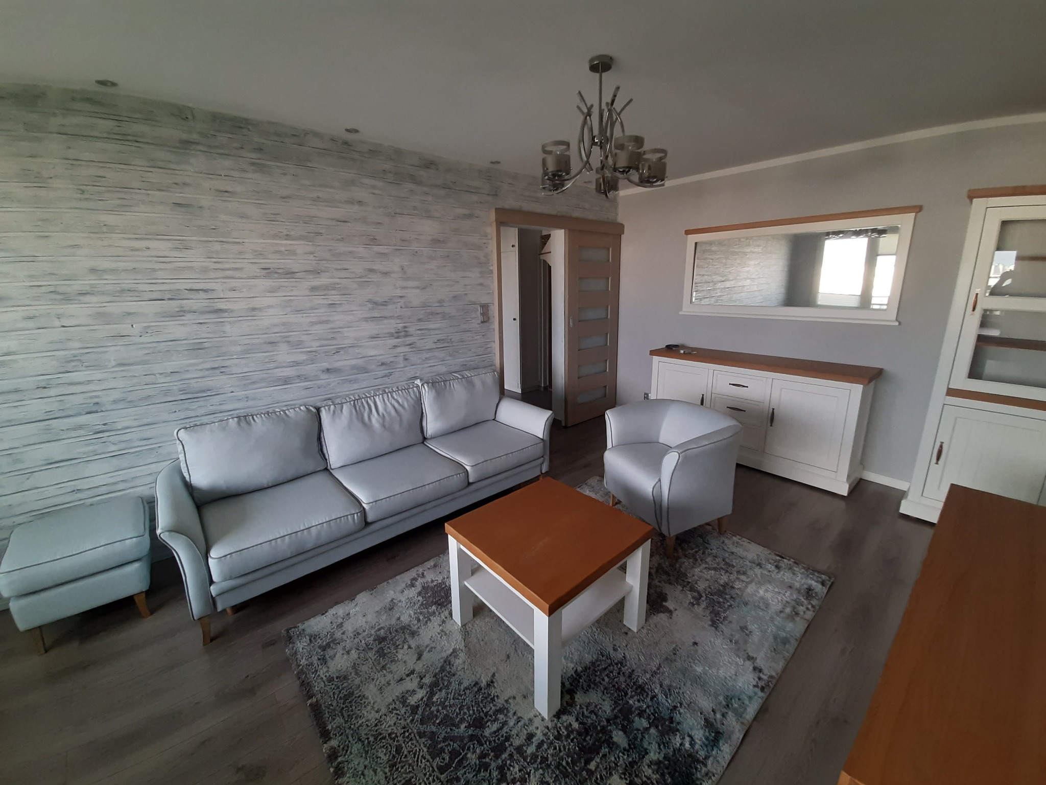 Nowoczesny salon z szarą sofą, fotelem i drewnianym stolikiem kawowym na wzorzystym dywanie; ściana wykończona w stylu imitującym bielone drewno.