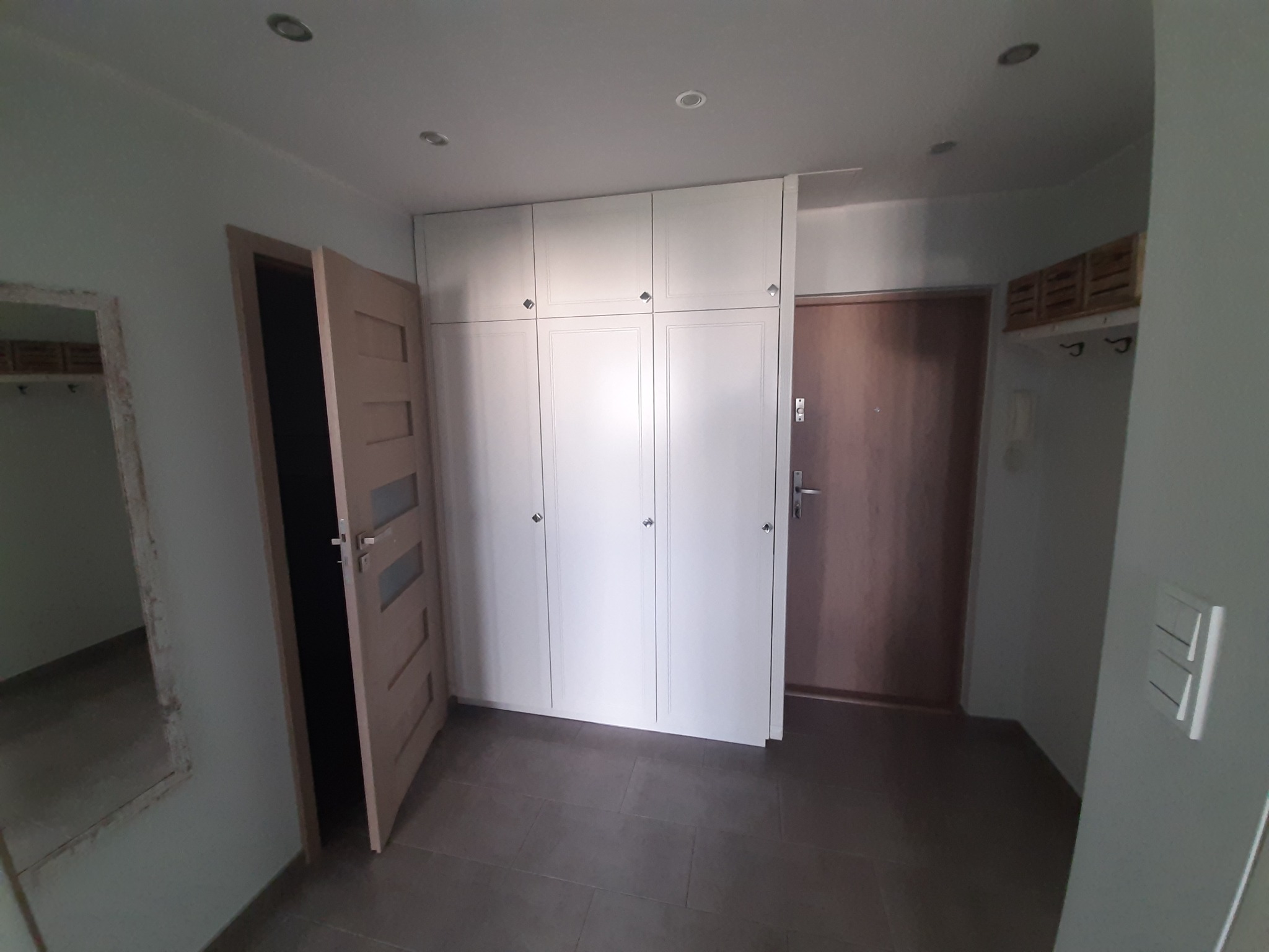 Przedpokój z zabudową szafy wnękowej z białymi frontami, brązową podłogą z płytek i otwartymi drzwiami do pokoju.