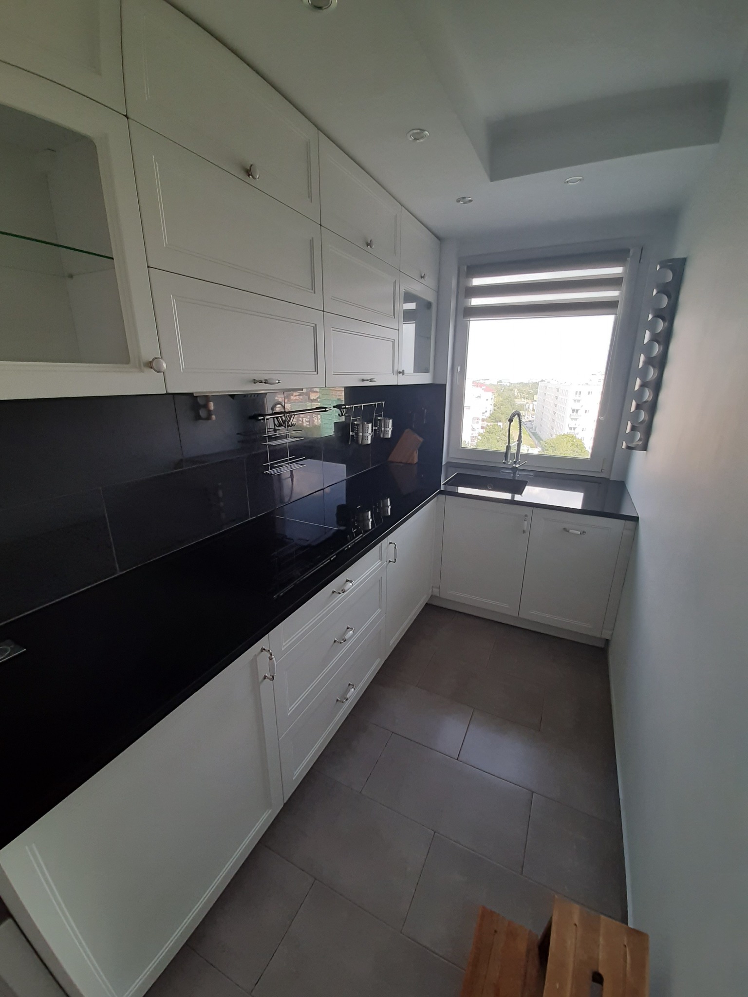 Nowoczesna, jasna kuchnia z białymi szafkami, czarnym blatem i szarą podłogą z płytek. Widoczne okno z roletami, metalowy ociekacz na naczynia i drewniany stołek na pierwszym planie.