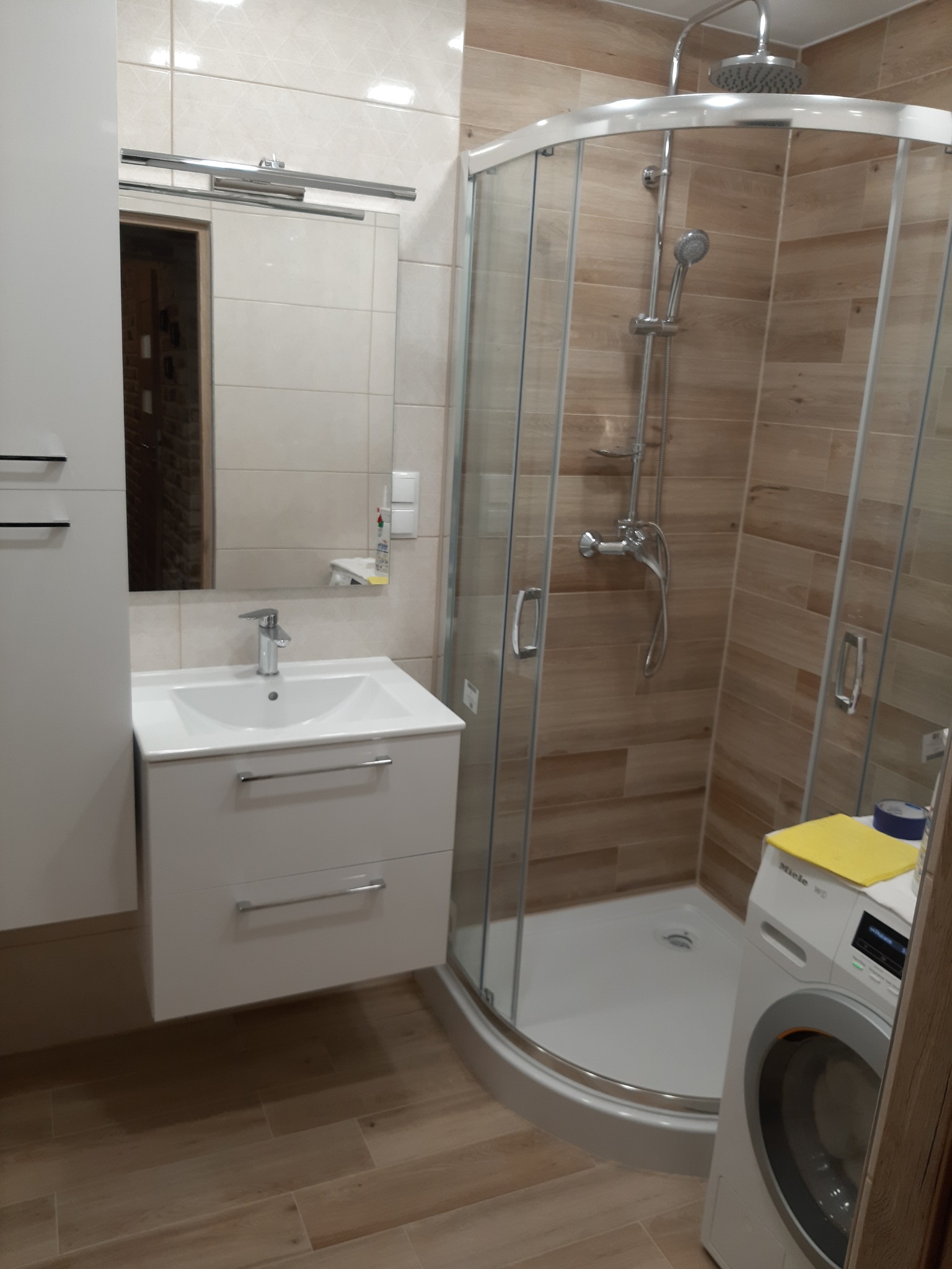 Nowoczesna łazienka z kabiną prysznicową, umywalką z szafką w kolorze białym, lustrem i pralką Miele. Płytki imitujące drewno na podłodze i ścianach. Ujęcie z perspektywy drzwi do łazienki.