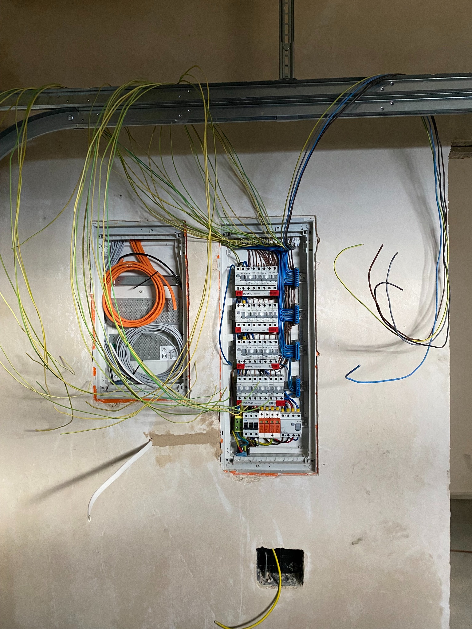Wewnętrzna instalacja elektryczna w trakcie montażu: widoczne dwie wnęki w surowej ścianie, jedna z rozdzielnicą z podłączonymi przewodami, druga z zwiniętymi kablami pomarańczowymi i szarymi,...
