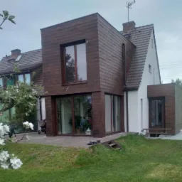 Nowoczesny dom jednorodzinny z elewacją łączącą biały tynk i ciemnobrązdy panel imitujący drewno, widok z ogrodu z kwitnącym drzewem owocowym.