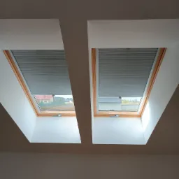 Dwa okna dachowe z drewnianą ramą i szarymi plisowanymi roletami wewnętrznymi, widok z wnętrza poddasza.