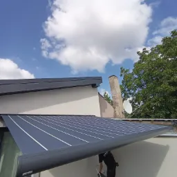 Montaż szarej markizy z białymi paskami na tarasie, pracownik na drabinie w trakcie instalacji, widok z dołu na tle błękitnego nieba z chmurami.