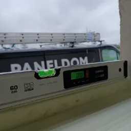 Poziomica cyfrowa wskazująca zero stopni, umieszczona na parapecie podczas montażu, w tle widoczny samochód dostawczy firmy z drabiną na dachu.