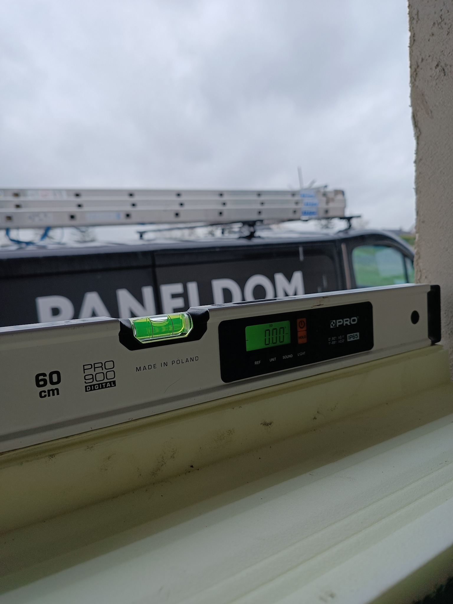 Poziomica cyfrowa wskazująca zero stopni, umieszczona na parapecie podczas montażu, w tle widoczny samochód dostawczy firmy z drabiną na dachu.