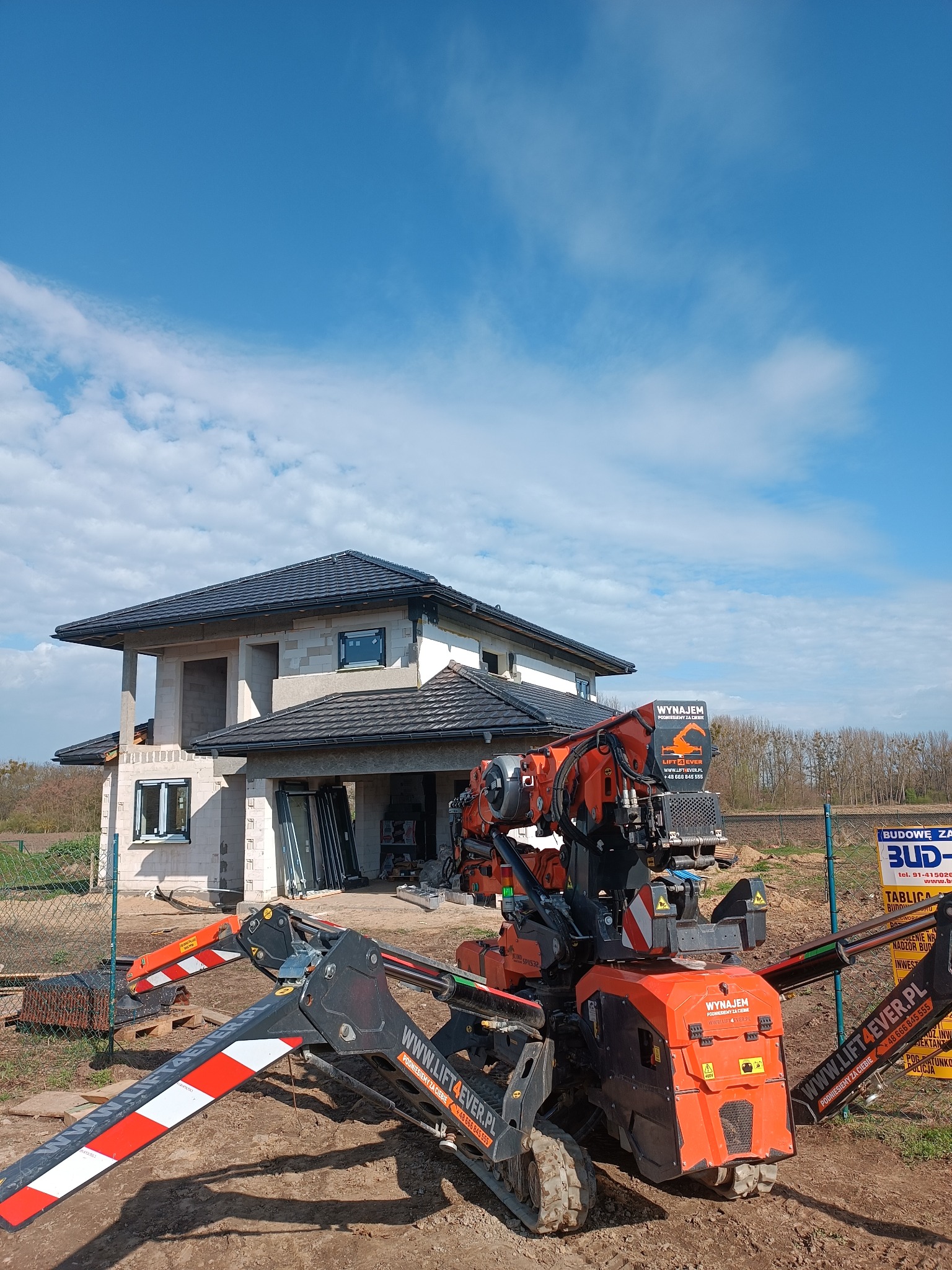 Montaż okien w nowym domu jednorodzinnym za pomocą mini-żurawia gąsienicowego, widoczne ramy okienne i elementy konstrukcyjne budynku w stanie surowym otwartym.