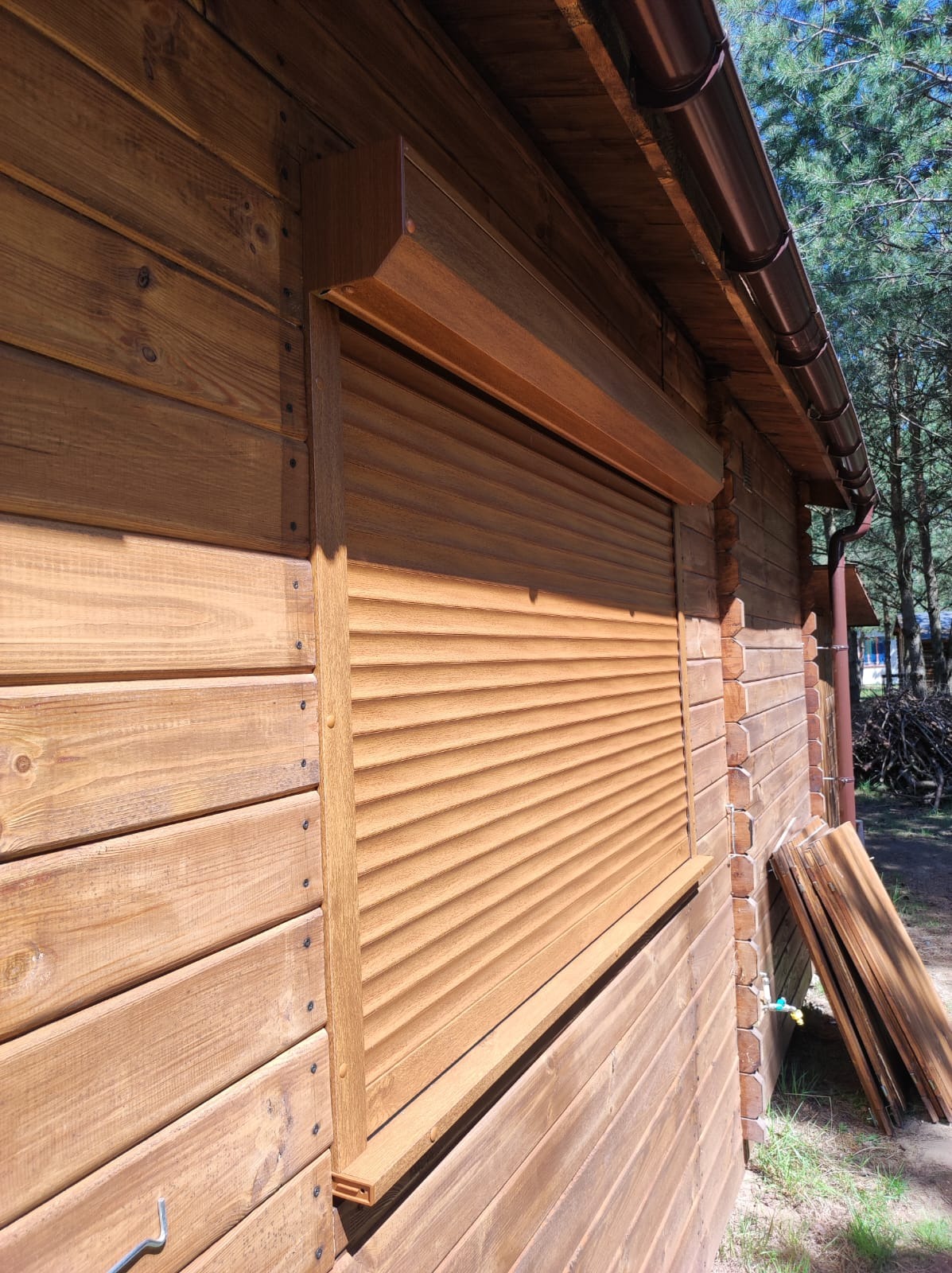 Drewniana elewacja domu z zamontowaną, brązową roletą zewnętrzną w pozycji zamkniętej, widok z boku na tle zieleni.