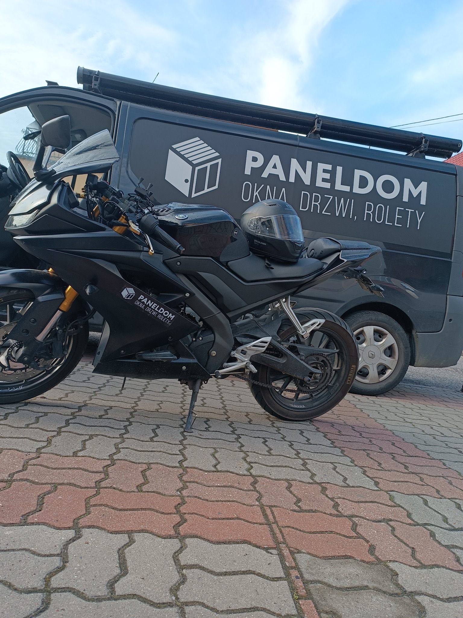 Czarny motocykl zaparkowany przed czarnym samochodem dostawczym z logo firmy Paneldom oferującej okna, drzwi i rolety.