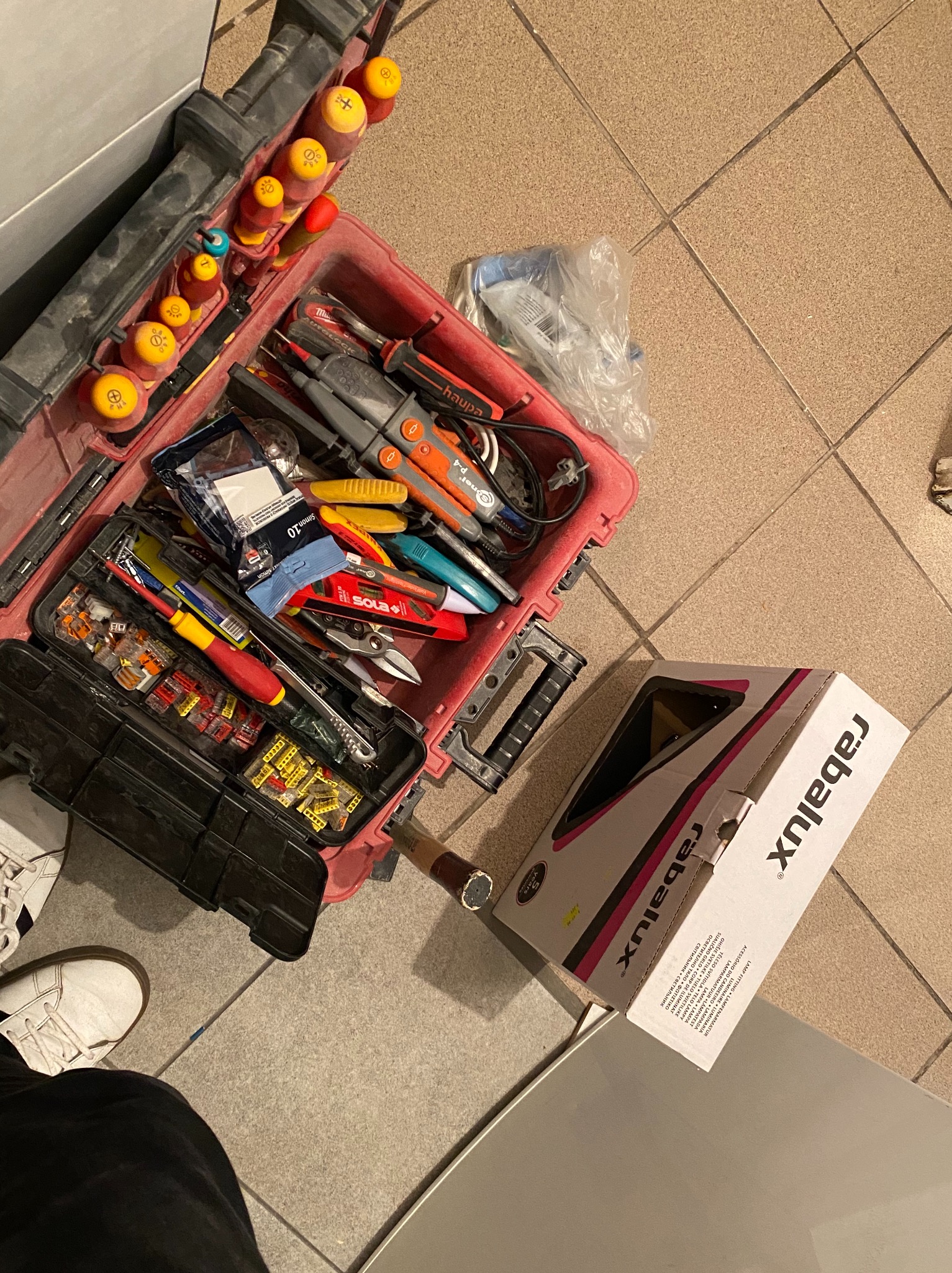 Otwarta skrzynka narzędziowa z różnorodnymi narzędziami elektrycznymi, złączkami i testerem napięcia, obok pudełko z lampą marki Rabalux, widok z góry na kafelkową podłogę.