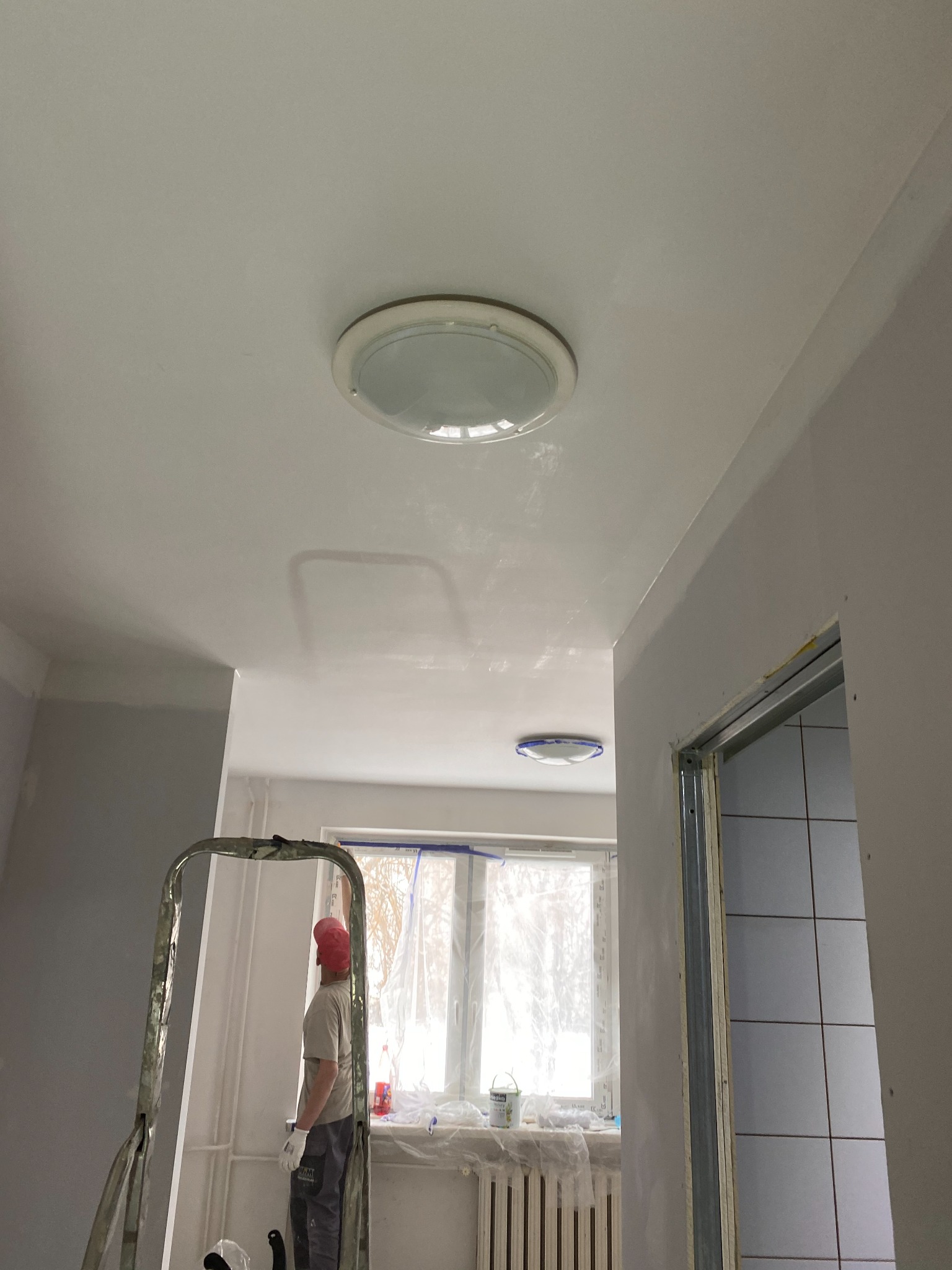 Pracownik w czerwonym kasku na drabinie podczas remontu sufitu z zamontowanym okrągłym oświetleniem w pomieszczeniu z oknem i grzejnikiem.