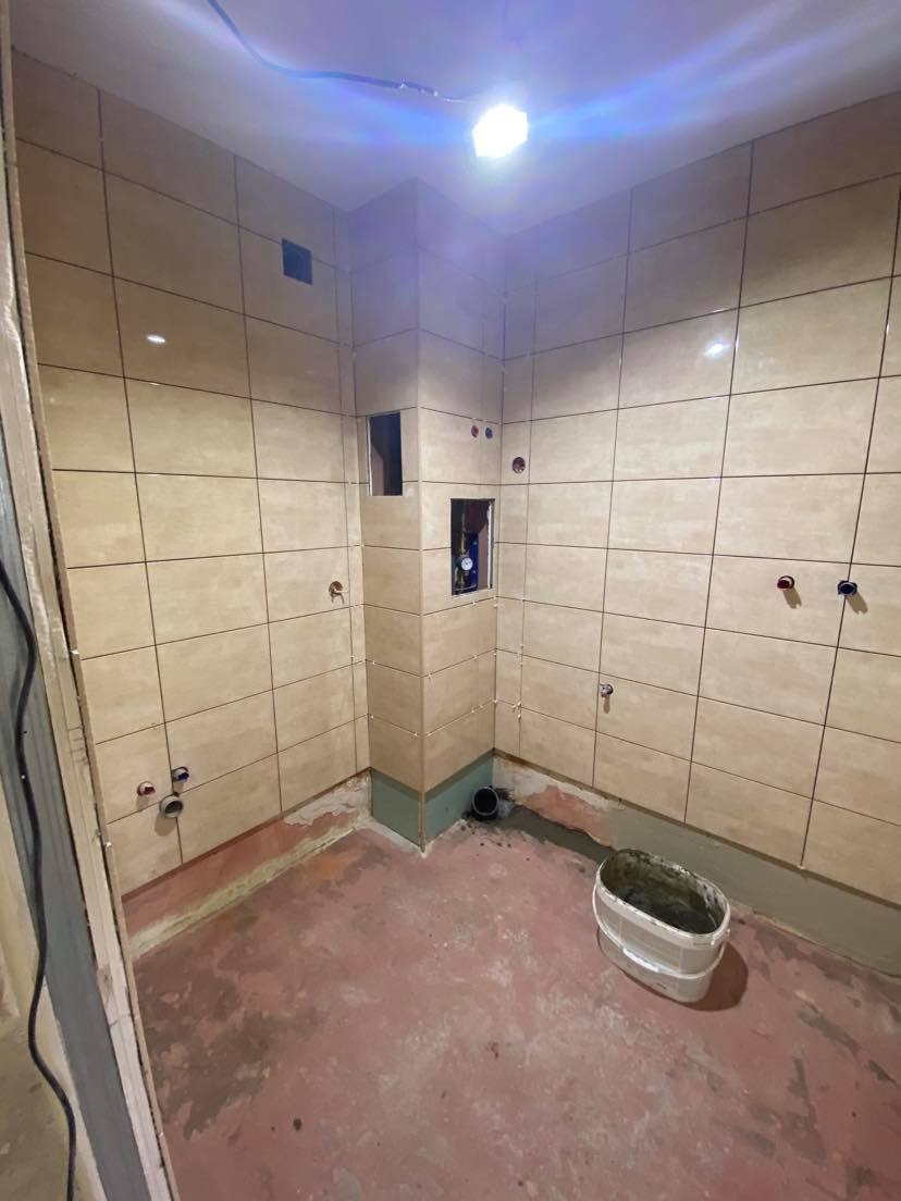 Wnętrze łazienki w trakcie remontu: ułożone płytki ceramiczne na ścianach, widoczne punkty przyłączy wodnych i kanalizacyjnych, otwory w ścianie, wiadro z zaprawą na podłodze.