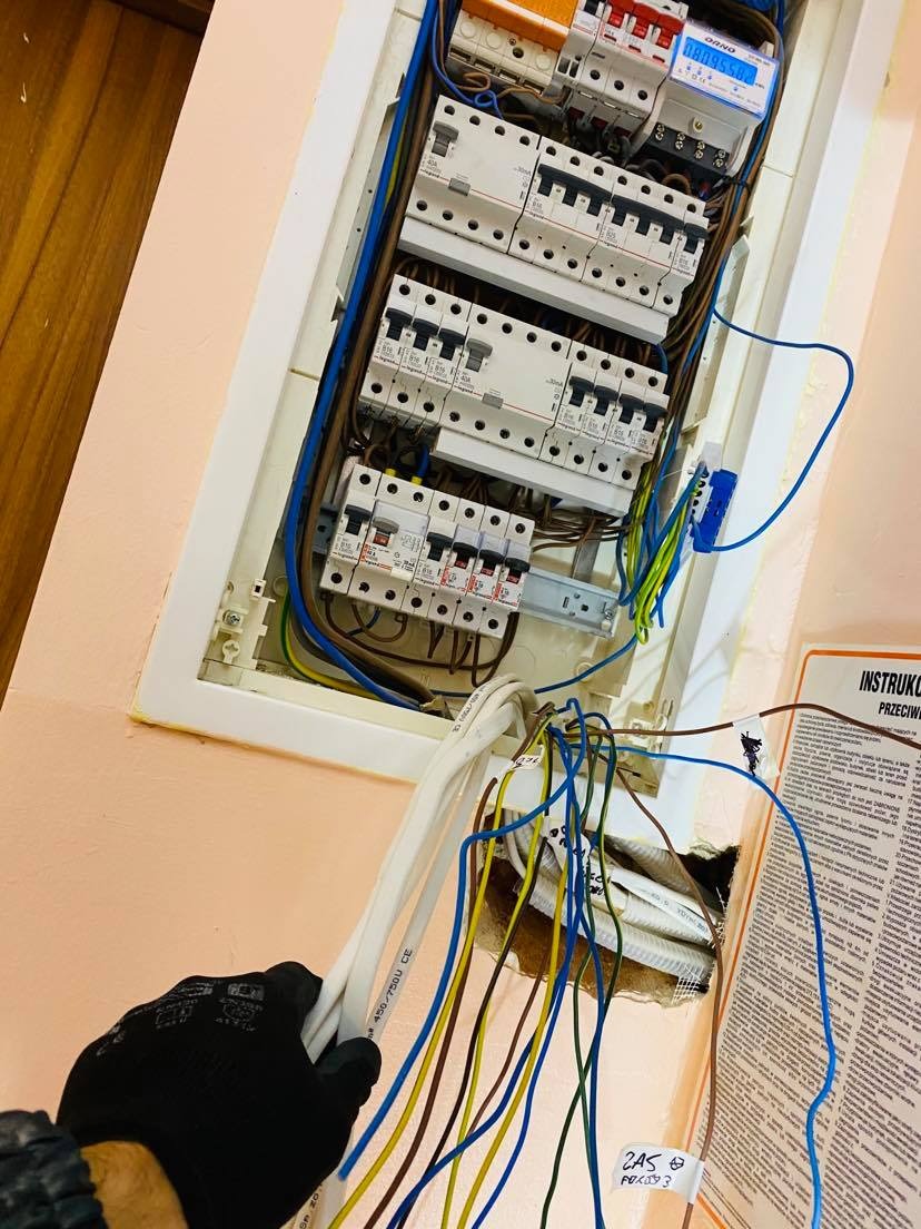 Elektryk w czarnych rękawicach podłącza przewody do skrzynki z bezpiecznikami, widoczne liczne kable w różnych kolorach i instrukcja bezpieczeństwa obok.