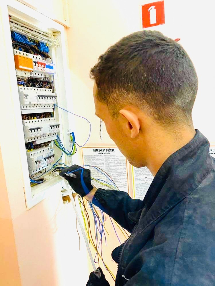 Elektryk w czarnych rękawicach podłącza przewody do skrzynki rozdzielczej z bezpiecznikami, widoczna instrukcja BHP na ścianie.