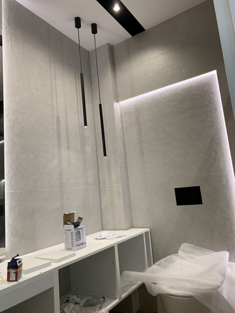 Nowoczesna łazienka w trakcie wykończenia z podwieszanymi lampami cylindrycznymi, listwą LED i częściowo zamontowanymi szafkami pod umywalkę.