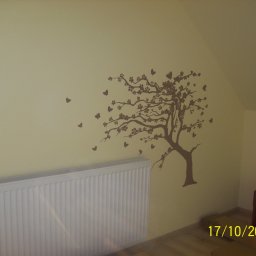 DORHAN PRACE BUDOWLANO - REMONTOWE KRYSTIAN LEWANDOWSKI - Ściana w pokoju na poddaszu pomalowana na kremowo, z naklejką w kształcie brązowego drzewa z motywem kwiatów i motyli, biały grzejnik pod ścianą.