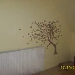 Ściana w pokoju na poddaszu pomalowana na kremowo, z naklejką w kształcie brązowego drzewa z motywem kwiatów i motyli, biały grzejnik pod ścianą.