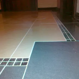 Błyszcząca podłoga wyłożona jasnobeżowymi i ciemnoszarymi płytkami z dekoracyjnym pasem mozaiki w przejściu, widoczne fugi i fragment listwy przypodłogowej.