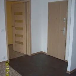 Korytarz po remoncie z dwiema parami drzwi w odcieniach drewna: jedne z przeszkleniami, drugie pełne, brązowa podłoga z płytek, fragment podłogi z paneli widoczny w lewym dolnym rogu.