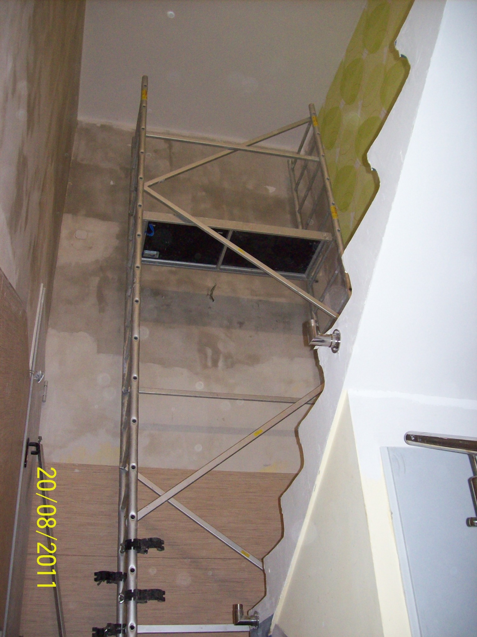 Wnętrze w trakcie prac przygotowawczych: metalowe rusztowanie na tle ściany z fragmentami starej tapety i surowego tynku, widoczny fragment drewnianej okładziny i drzwi.