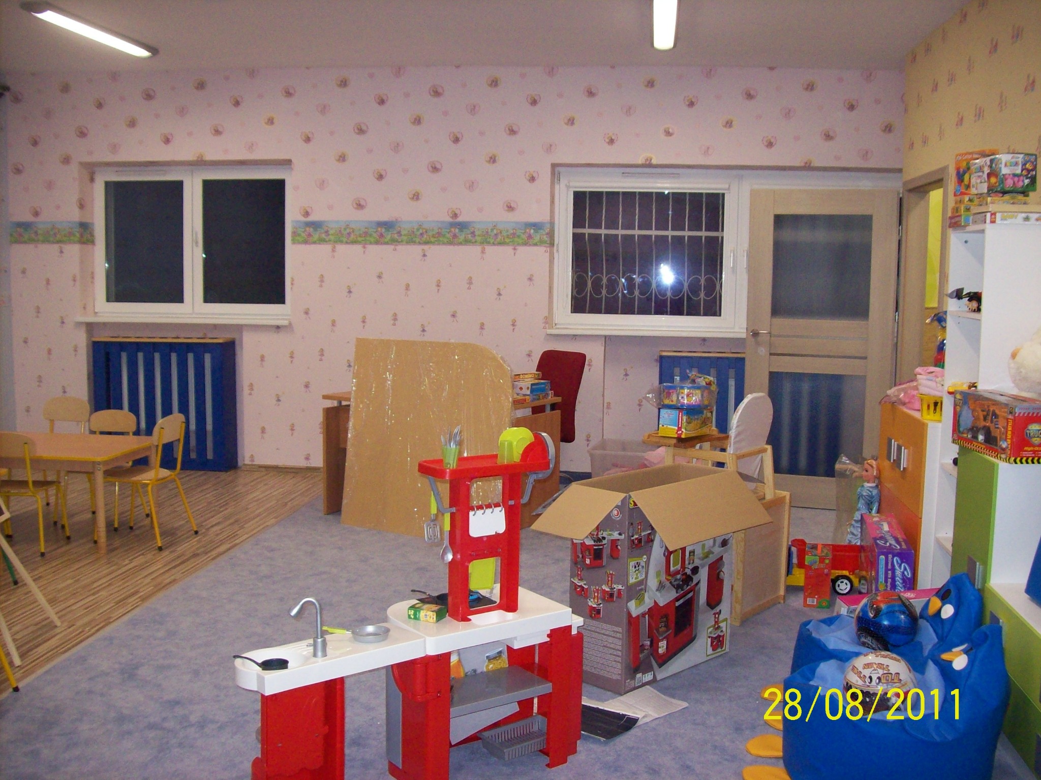 Pokój dziecięcy z jasnymi ścianami w kwiatki, wyposażony w drewniane meble, stolik z krzesłami, plastikową kuchnię zabawkową, tekturowy domek i niebieski fotel puf. Widoczne okna z kratami...