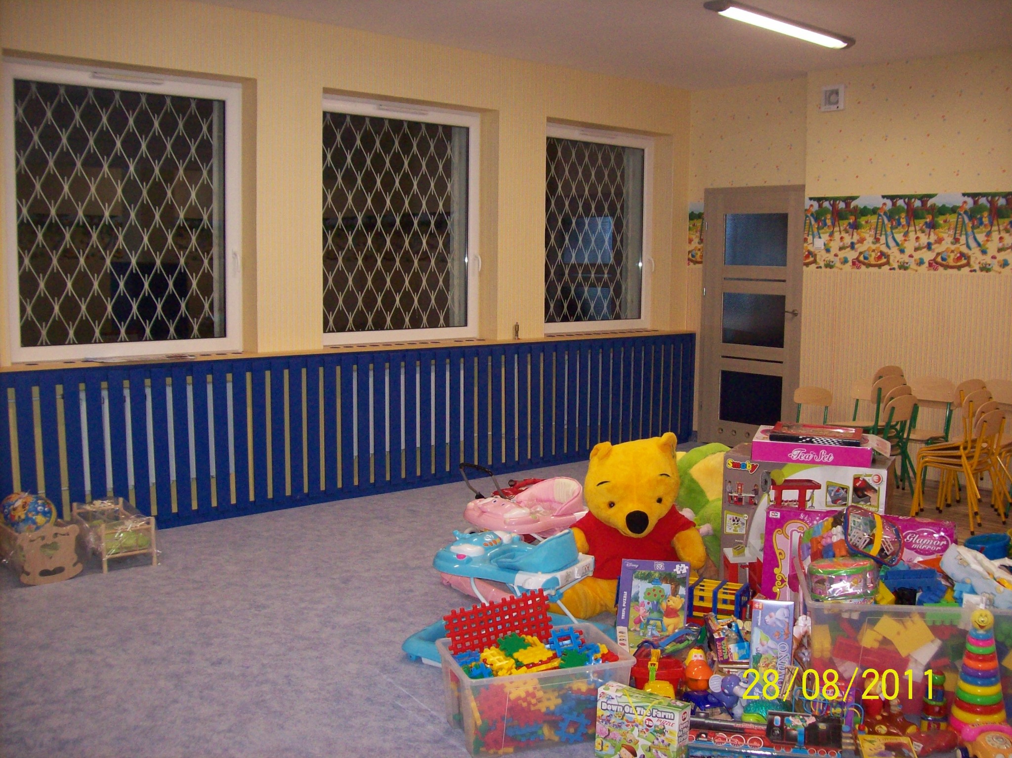 Wnętrze pokoju dziecięcego z trzema oknami zabezpieczonymi kratami, niebieską barierką pod oknami i tapetą z motywem dziecięcym. Na podłodze zgromadzone różnorodne zabawki, w tym pluszowy Kubuś...