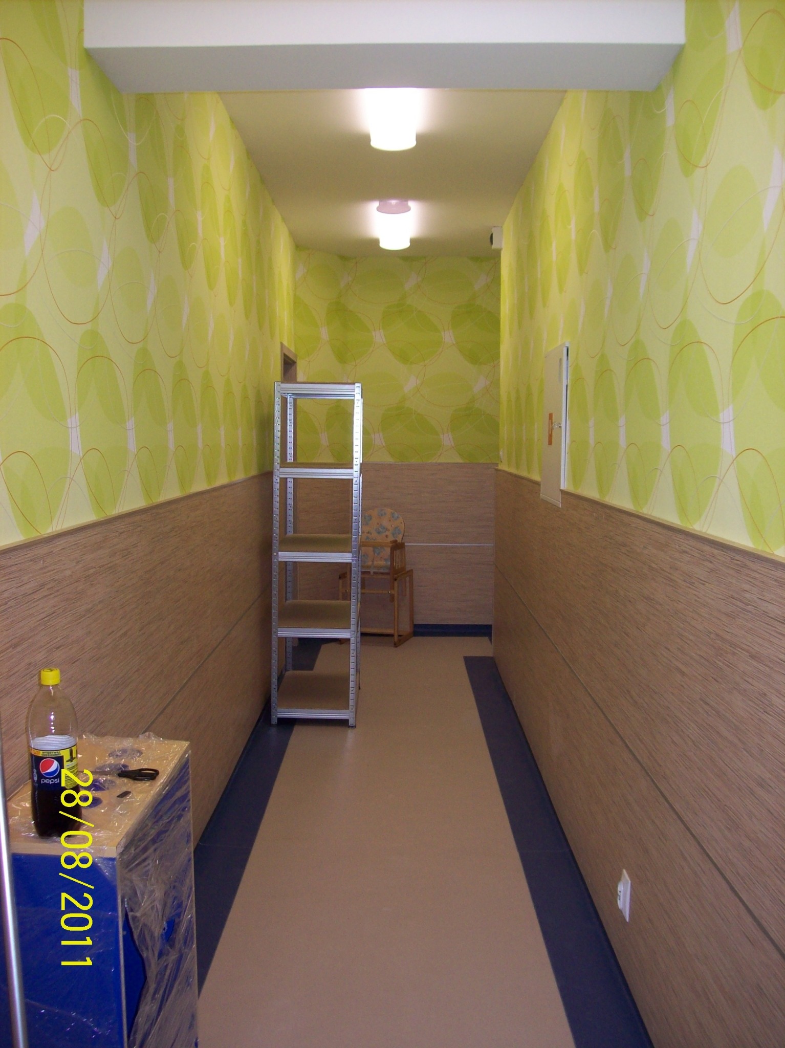 Korytarz z zieloną tapetą w okręgi i drewnianą boazerią, metalowy regał z pustymi półkami, butelka napoju na niebieskiej szafce w folii, podłoga w odcieniach beżu i granatu.