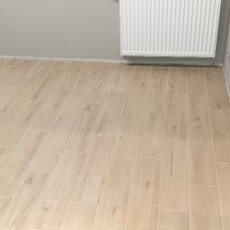 Koma-Bud s.c. - Ułożona podłoga z płytek imitujących drewno w jasnym odcieniu, widoczny grzejnik na ścianie i szara ściana, świeżo po fugowaniu.