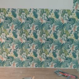 Koma-Bud s.c. - Ściana i róg pokoju pokryte tapetą w tropikalne liście, widoczne narzędzia i fragment podłogi w trakcie prac wykończeniowych.